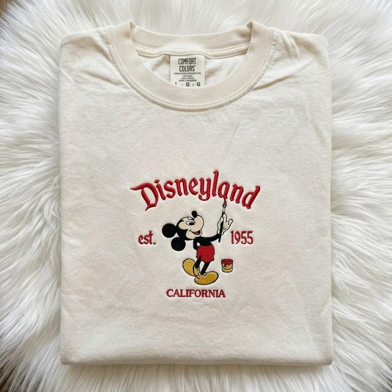 May include: A cream-coloured short-sleeve t-shirt featuring an embroidered design of Mickey Mouse holding a paintbrush. The text reads Disneyland, est. 1955, and CALIFORNIA in red thread. The shirt has a Comfort Colors brand label at the inner neckline.