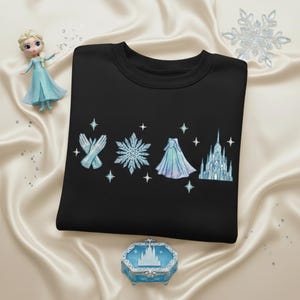 May include: Black sweatshirt with embroidered light blue gloves, snowflake, dress, and castle designs. A small figurine and a decorative box with a castle design are also present. The items are arranged on a satin surface.