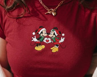 May include: A dark red t-shirt featuring a central embroidered graphic of two cartoon mice wearing red and white Santa hats and green scarves. The mice are facing each other, forming a heart shape with their hands. A gold chain necklace is worn.