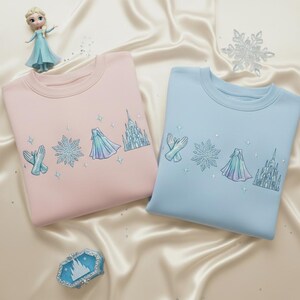 May include: Two crewneck sweatshirts, one light pink and one light blue, featuring embroidered designs of a snowflake, gloves, a cape, and a castle. A small doll and decorative snowflake are also visible.