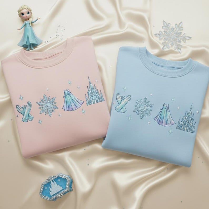 May include: Two crewneck sweatshirts, one light pink and one light blue, featuring embroidered designs of a snowflake, gloves, a cape, and a castle. A small doll and decorative snowflake are also visible.