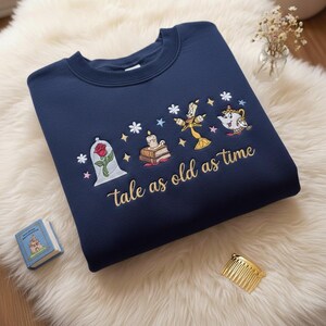 Puede incluir: Sudadera azul marino con diseño bordado que presenta una rosa en una cúpula de cristal, una vela, libros, Lumiere y la Sra. Potts. La frase "tale as old as time" está bordada en dorado. La sudadera está sobre una superficie blanca y esponjosa.