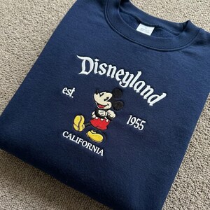 May include: A navy blue crewneck sweatshirt featuring white embroidered text that reads Disneyland est. 1955 CALIFORNIA. Centered is an embroidered graphic of Mickey Mouse wearing red shorts and yellow shoes, with a white interior neck label visible.