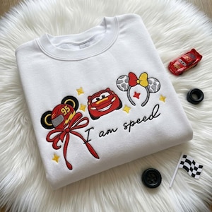 May include: A white crewneck sweatshirt featuring embroidered designs of a red race car, a racing helmet with the number 95, and mouse ears with a bow. The text "I am speed" is embroidered in black script below the colorful car graphics.