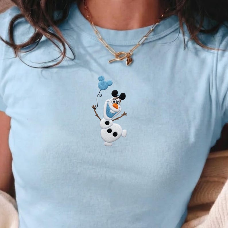 May include: Light blue t-shirt featuring an embroidered Olaf snowman holding a blue Mickey Mouse balloon. The shirt has a crew neck and short sleeves. A gold necklace is worn around the neck.
