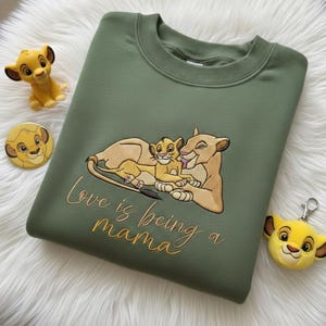 May include: An olive green sweatshirt with an embroidered design of a lioness and cub, with the text "Love is being a mama" in gold script. The image also includes a Simba figurine, a Simba button, and a Simba keychain.