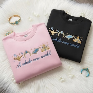 Puede incluir: Dos sudaderas, una rosa y otra negra, bordadas con una lámpara dorada, una gema azul, una alfombra mágica y un palacio. El texto "A whole new world" está bordado en azul en ambas sudaderas. También se ven pulseras doradas y turquesas.