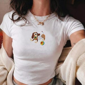 May include: White t-shirt featuring a cartoon dachshund dog with a spring body, a green collar, and a yellow and red ball. The dog is brown, tan, and white. The shirt has short sleeves and a crew neck.