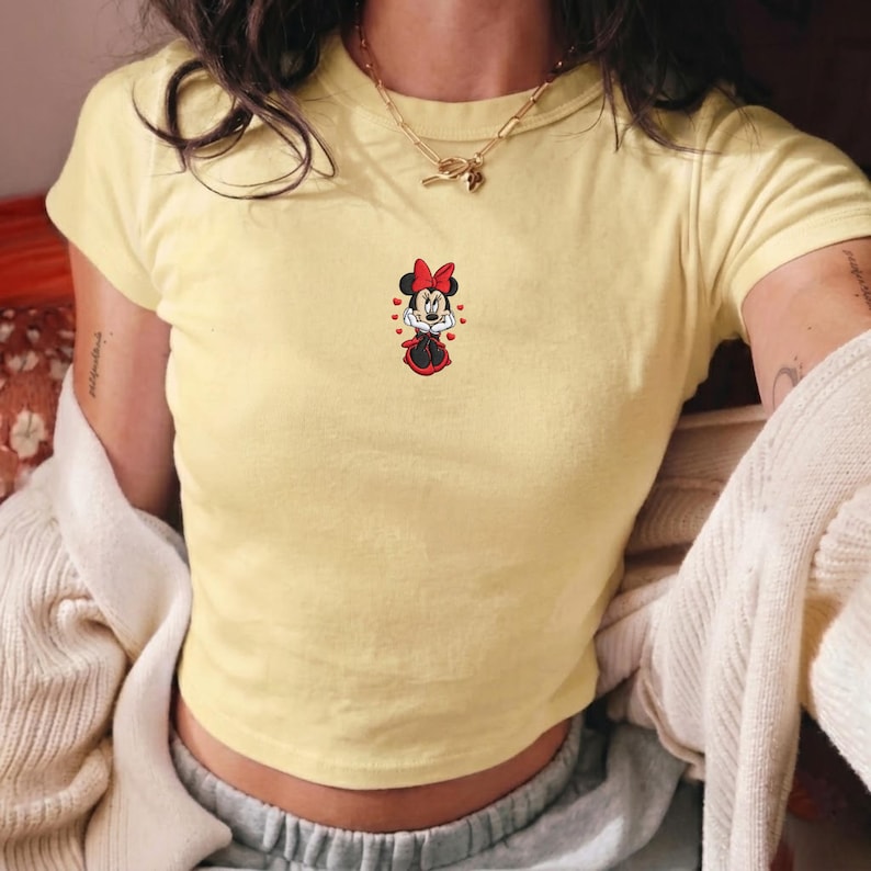 May include: A light yellow crop top featuring a small embroidered Minnie Mouse design. The design includes a red bow, dress, and heart accents. The shirt has short sleeves and a round neckline. A gold necklace is visible.