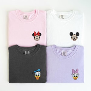 Könnte beinhalten: Vier gefaltete T-Shirts in Rosa, Weiß, Anthrazit und Lavendel. Jedes Shirt zeigt einen anderen gestickten Disney-Charakter: Minnie Mouse, Mickey Mouse, Donald Duck und Daisy Duck. Die Shirts sind von der Marke Comfort Colors.