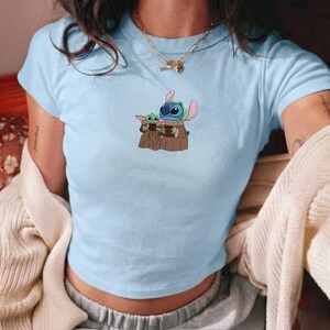 Op de afbeelding: Lichtblauw crop top met een afbeelding van Stitch en Grogu uit Star Wars. De figuren houden bruine voorwerpen vast. Het shirt heeft korte mouwen en een ronde hals. Een casual kledingstuk.