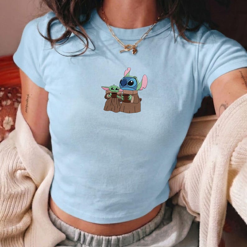 Op de afbeelding: Lichtblauw crop top met een afbeelding van Stitch en Grogu uit Star Wars. De figuren houden bruine voorwerpen vast. Het shirt heeft korte mouwen en een ronde hals. Een casual kledingstuk.
