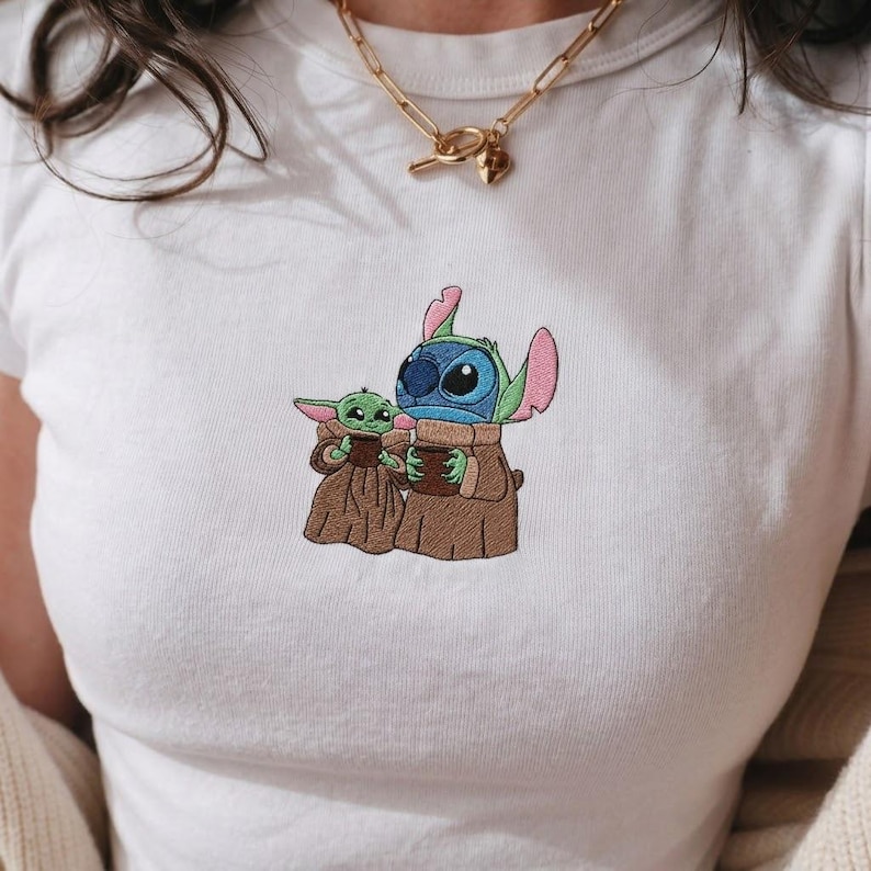 Op de afbeelding: Wit T-shirt met een geborduurd ontwerp van Stitch en Grogu, beiden in bruine gewaden, die een bruine beker vasthouden. Stitch is blauw met grote oren en Grogu is groen. Een gouden ketting is zichtbaar.
