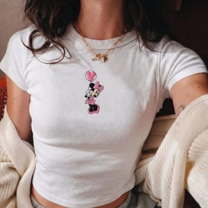 May include: White crop top featuring a Minnie Mouse graphic holding a pink balloon. The cartoon character is wearing a pink dress, bow, and shoes. The shirt has short sleeves and a crew neck.