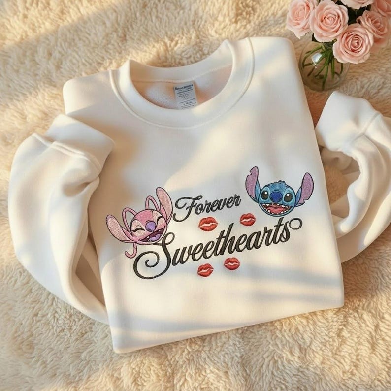 May include: White sweatshirt featuring the phrase "Forever Sweethearts" with cartoon characters and red lip graphics. The sweatshirt has long sleeves and a crew neck. The design includes a pink and blue cartoon character.