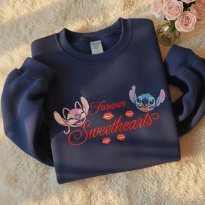 May include: Navy blue sweatshirt featuring embroidered cartoon characters and the text "Forever Sweethearts" in red. The design includes two characters and red lip prints. The sweatshirt is a casual wear item.