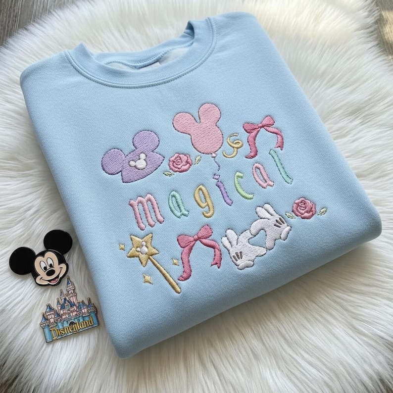 May include: A light blue sweatshirt with embroidered details. The design includes Mickey Mouse ears, bows, roses, a wand, and the word "magical" in colorful letters. A Mickey Mouse pin and a Disneyland pin are next to the sweatshirt.