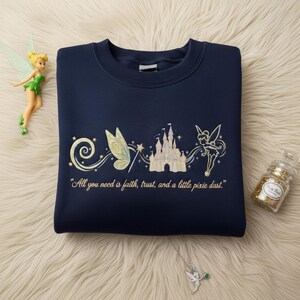 May include: Navy blue sweatshirt with embroidered fairy, castle, and text "All you need is faith, trust, and a little pixie dust." A fairy figurine and a glitter jar are also visible.