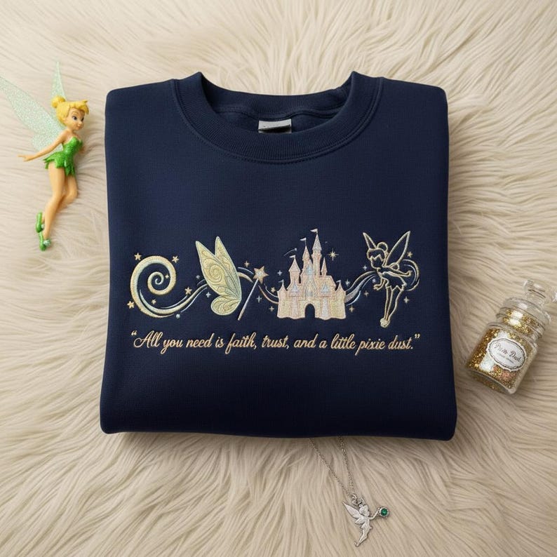 May include: Navy blue sweatshirt with embroidered fairy, castle, and text "All you need is faith, trust, and a little pixie dust." A fairy figurine and a glitter jar are also visible.