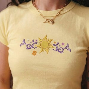 May include: A pale yellow short-sleeved t-shirt featuring a central embroidered design of a golden sun surrounded by purple swirling patterns, small orange and white flowers, and crescent moon shapes, paired with a gold-toned chain necklace.