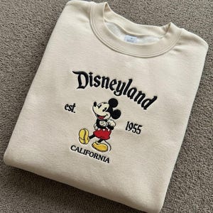 May include: A beige crewneck sweatshirt featuring black embroidered text that reads Disneyland est. 1955 CALIFORNIA. Centered is an embroidered graphic of Mickey Mouse wearing red shorts and yellow shoes, positioned between the text elements.