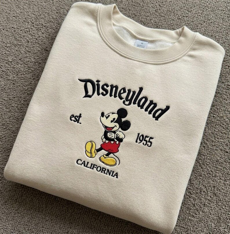 May include: A beige crewneck sweatshirt featuring black embroidered text that reads Disneyland est. 1955 CALIFORNIA. Centered is an embroidered graphic of Mickey Mouse wearing red shorts and yellow shoes, positioned between the text elements.