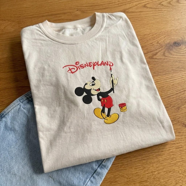 Pu&ograve; includere: T-shirt color crema con grafica Disneyland. Il design include Topolino che tiene un pennello, con la scritta "Disneyland" in rosso. La maglietta &egrave; abbinata a jeans di denim blu chiaro.