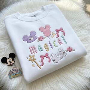 May include: White sweatshirt with colorful embroidered design. Features the word "magical" in pastel colors, Mickey Mouse head shapes, bows, roses, a wand, and Mickey Mouse gloves. A Mickey Mouse pin and Disneyland pin are next to the sweatshirt.