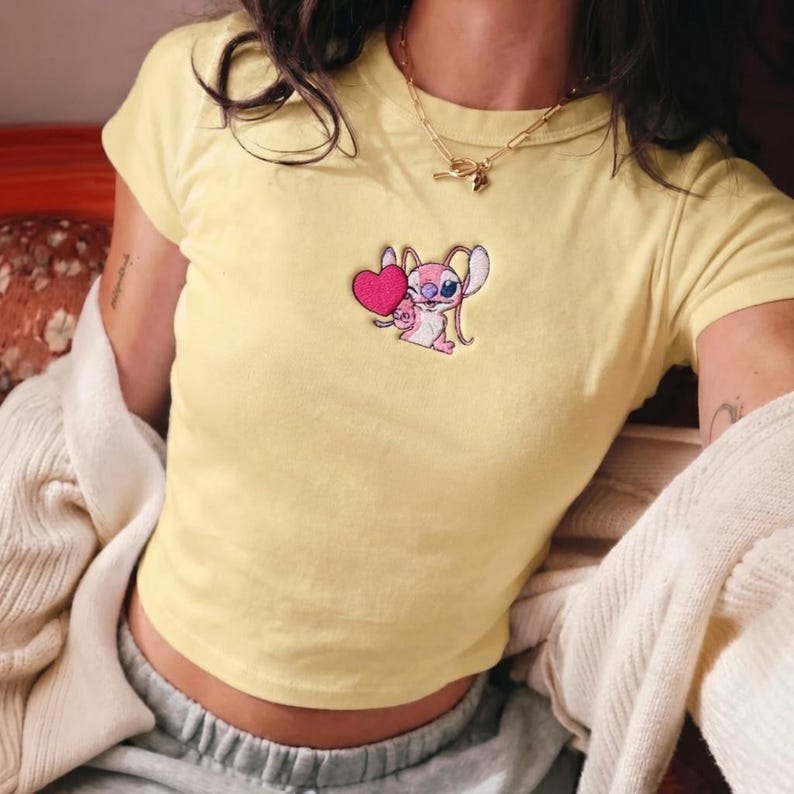 May include: Yellow crop top with a small embroidered design of a cartoon character holding a pink heart. The shirt has short sleeves and a crew neck. A gold necklace is visible.