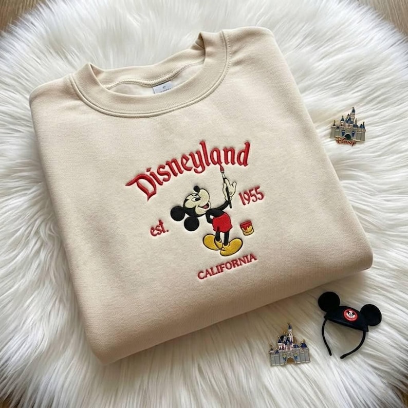 May include: A beige crewneck sweatshirt featuring an embroidered graphic of Mickey Mouse holding a paintbrush. The text reads Disneyland, est. 1955, and California in red lettering. The design includes a small paint bucket and a castle pin.