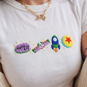 May include: White t-shirt with embroidered designs from the animated film Toy Story. The shirt features the word "ANDY" in purple, a purple and green space ranger, a blue rocket, and a yellow ball with a red star.