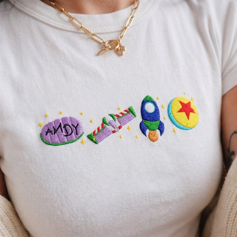 May include: White t-shirt with embroidered designs from the animated film Toy Story. The shirt features the word "ANDY" in purple, a purple and green space ranger, a blue rocket, and a yellow ball with a red star.