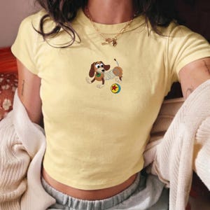 May include: A light yellow crop top featuring an embroidered cartoon dachshund dog with a spring body, a green collar, and a red and yellow ball. The dog is in a playful pose. The shirt has short sleeves and a round neckline.