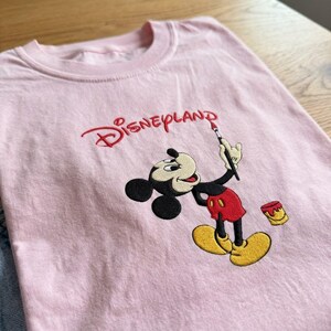 Pu&ograve; includere: T-shirt rosa chiaro con la scritta "Disneyland" ricamata in rosso. La maglietta presenta un'immagine ricamata di Topolino che tiene un pennello, con pantaloncini rossi e scarpe gialle.