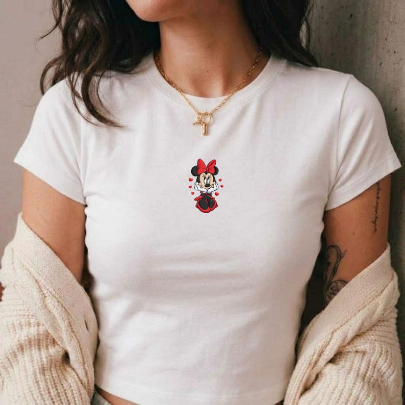 May include: White t-shirt featuring a Minnie Mouse graphic. The cartoon character is depicted in a red dress and bow, with heart accents. The shirt has short sleeves and a crew neck.