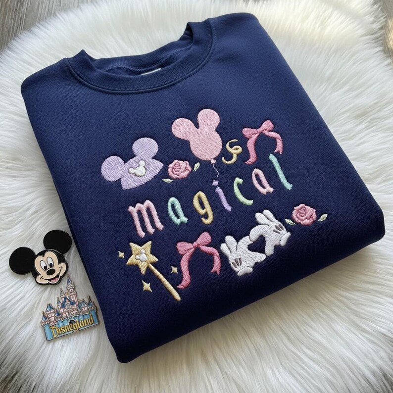 May include: Navy blue sweatshirt with embroidered "magical" design. Features Mickey Mouse head, bows, roses, a wand, and Mickey Mouse gloves. A Mickey Mouse pin and Disneyland pin are next to the sweatshirt.