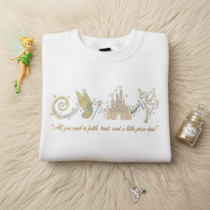 May include: White sweatshirt with embroidered design featuring a fairy, castle, and the text "All you need is faith, trust, and a little pixie dust." A fairy figurine, a necklace, and a jar of glitter are also in the image.