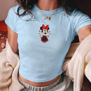 May include: A light blue short-sleeved t-shirt featuring a centered graphic of Minnie Mouse wearing a red bow and dress, surrounded by small red hearts. The character has her hands pressed against her cheeks in a pose with a neutral expression.