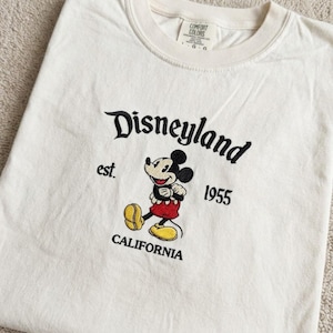 May include: An off-white t-shirt featuring black embroidered text that reads Disneyland, est. 1955, and CALIFORNIA. Centered is an embroidered Mickey Mouse character wearing red shorts and yellow shoes. The inner neck label reads Comfort Colors.