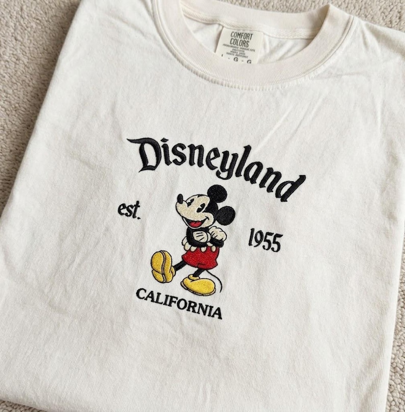 May include: An off-white t-shirt featuring black embroidered text that reads Disneyland, est. 1955, and CALIFORNIA. Centered is an embroidered Mickey Mouse character wearing red shorts and yellow shoes. The inner neck label reads Comfort Colors.