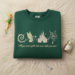 May include: A dark green sweatshirt with an embroidered design featuring a fairy, castle, and the text "All you need is faith, trust, and a little pixie dust." A fairy figurine and a jar of glitter are also visible.