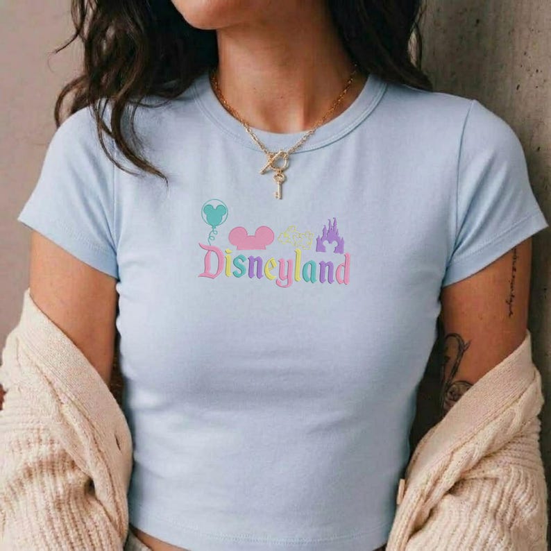 May include: Light blue t-shirt with a pastel Disneyland graphic. The design includes a heart-shaped balloon, Mickey Mouse ears, a castle, and the word "Disneyland" in a colorful, whimsical font. The shirt has short sleeves and a crew neck.