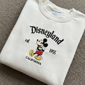 May include: A cream-coloured crewneck sweatshirt featuring an embroidered graphic of Mickey Mouse. The text reads Disneyland, est. 1955, and California. The embroidery uses black, red, and yellow thread to depict the character and lettering.