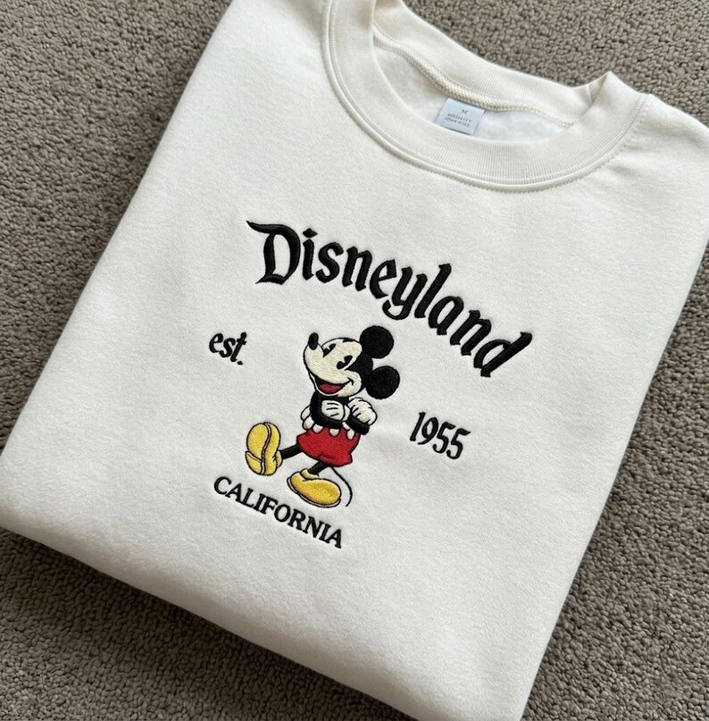 May include: A cream-coloured crewneck sweatshirt featuring an embroidered graphic of Mickey Mouse. The text reads Disneyland, est. 1955, and California. The embroidery uses black, red, and yellow thread to depict the character and lettering.