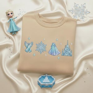 May include: A tan crewneck sweatshirt with embroidered light blue and white snowflake, gloves, dress, and castle designs. A small doll and a decorative box with a castle design are also in the image.