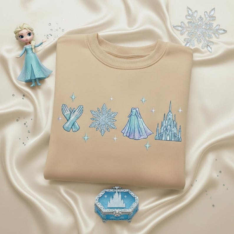 May include: A tan crewneck sweatshirt with embroidered light blue and white snowflake, gloves, dress, and castle designs. A small doll and a decorative box with a castle design are also in the image.