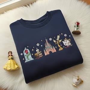 Puede incluir: Sudadera azul marino con un diseño bordado de personajes de La Bella y la Bestia. El diseño incluye una rosa en una campana de cristal, un castillo y otros personajes. Una pequeña figura de Bella está en la esquina inferior izquierda.