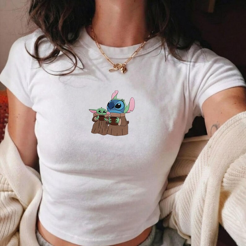 Op de afbeelding: Wit T-shirt met een kleurrijke afbeelding van Stitch en Grogu van Disney. De stripfiguren zijn afgebeeld terwijl ze elkaar omarmen. Het shirt heeft een ronde hals en korte mouwen.