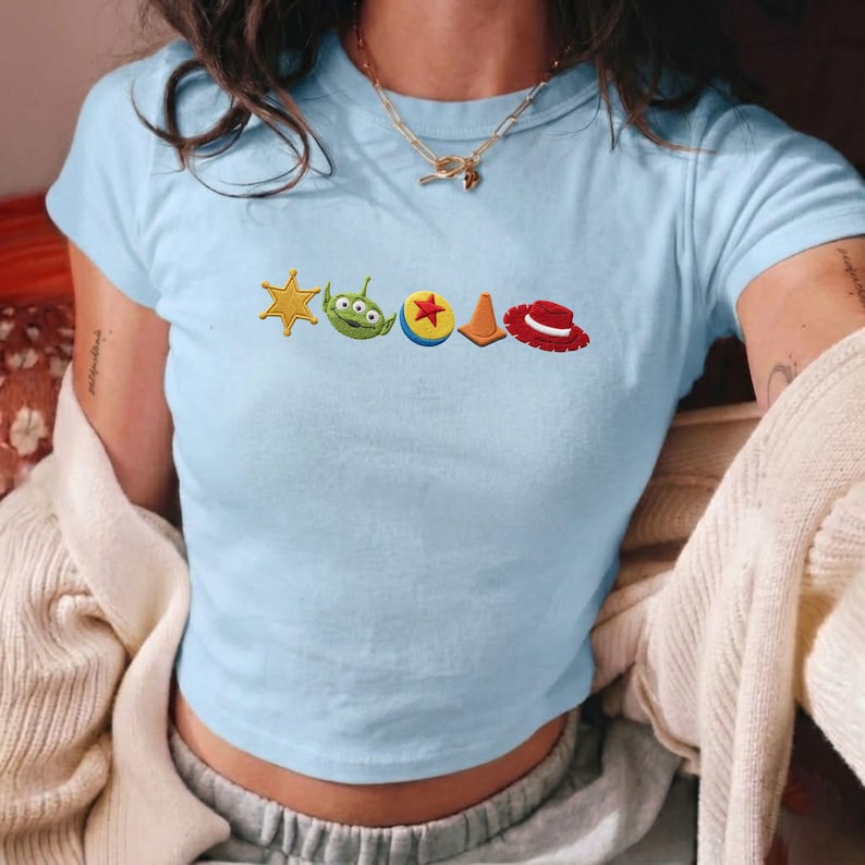 K&ouml;nnte beinhalten: Ein hellblaues T-Shirt mit kurzen &Auml;rmeln und einer horizontalen Reihe gestickter Symbole, darunter ein gelber Sheriffstern, ein gr&uuml;ner Alienkopf, ein blau-gelber Ball mit rotem Stern, ein orangefarbener Verkehrskegel und ein roter Cowboyhut.