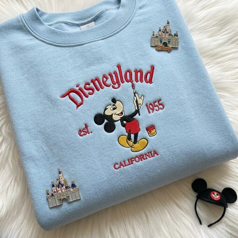 May include: A light blue crewneck sweatshirt featuring embroidered text that reads Disneyland est. 1955 CALIFORNIA with a graphic of Mickey Mouse holding a paintbrush. Two silver-toned castle pins are attached, and a black mouse ear headband is nearby.